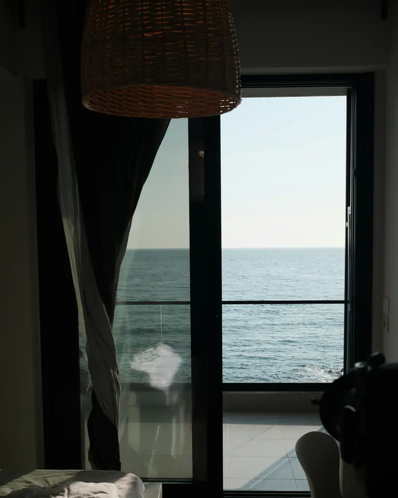 Baie vitrée sur la mer et l’horizon, rideau sombre et lit défait au premier plan